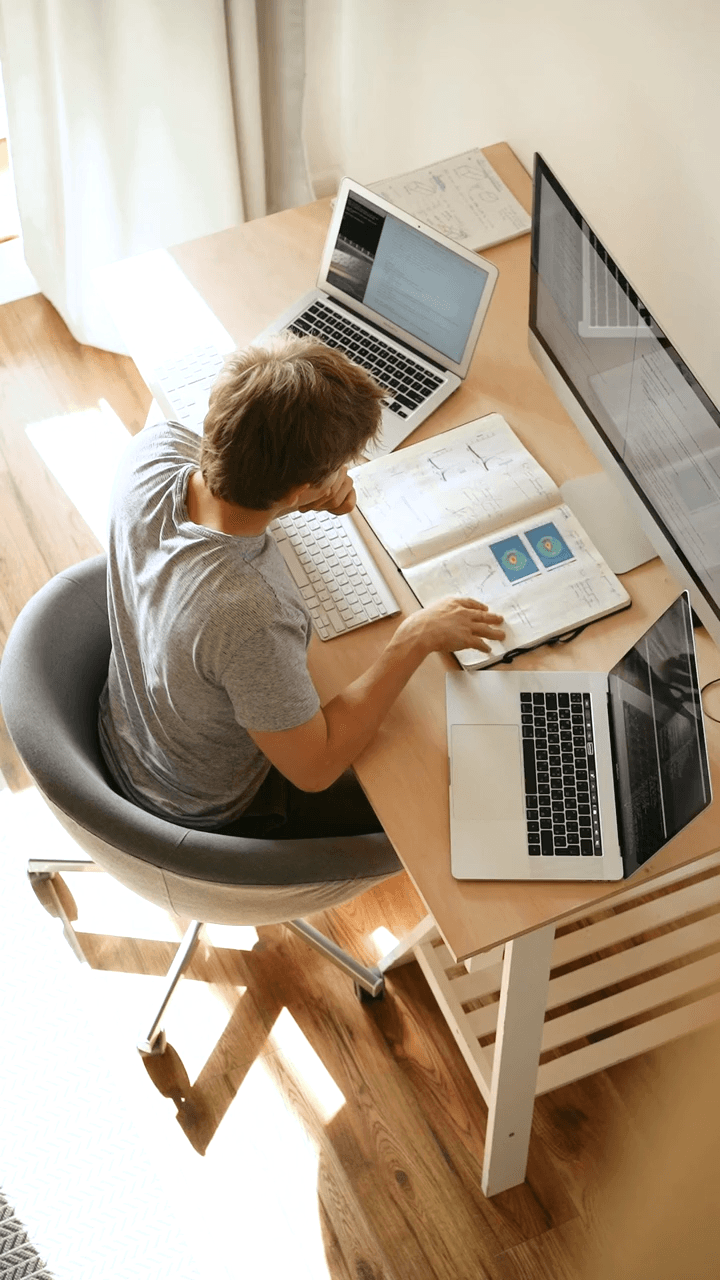 A person working at a desk with two laptops and an open notebook.